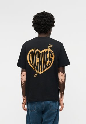 Person mit dunklen lockigen Haaren und Vitiligo an den Armen trägt ein schwarzes T-Shirt mit großem gelbem Herz und Pfeil-Grafik und dem Schriftzug "Dickies" auf der Rückseite.
