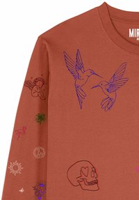 Chemise à manches longues de couleur rouille avec des colibris bleus sur la poitrine et des imprimés sur les manches comprenant un chérubin, un trèfle à quatre feuilles, un flamant rose, un signe de paix, un crâne et un soleil.