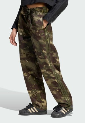Person trägt lockere Hose mit Tarnmuster und grünen Streifen, schwarze Sneaker mit weißen Streifen und ein schwarzes Langarmshirt mit Manschettenknopf.