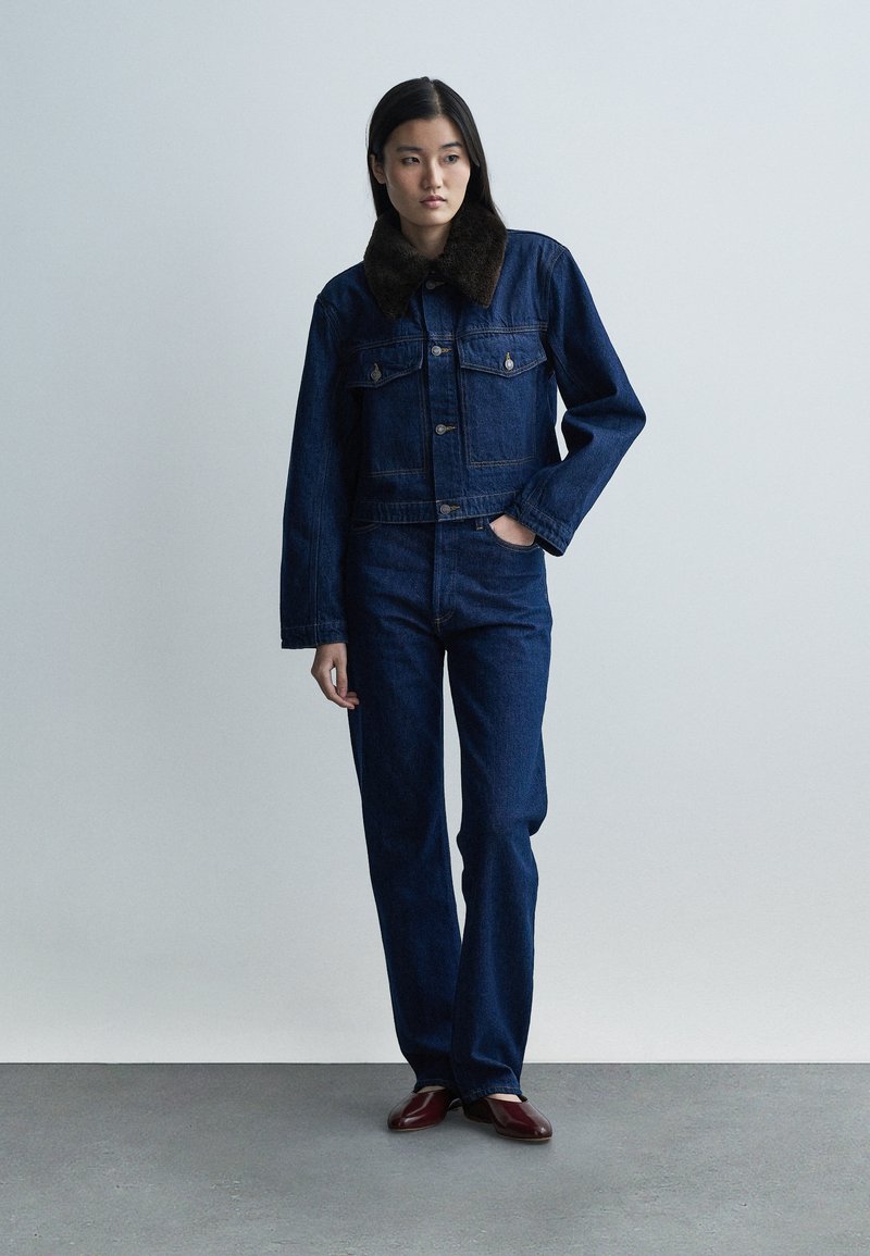 Denim jacket with a dark fur collar, featuring chest pockets and a button closure, paired with flared dark blue denim jeans and burgundy shoes.