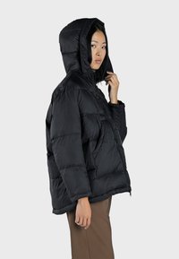 Doudoune noire avec capuche, fermeture éclair et texture matelassée. Présente une coupe ample et des poches latérales.