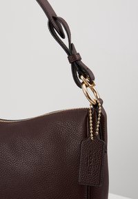 Sac à main en cuir grainé marron foncé avec fermeture éclair dorée, étiquette chaîne dorée et bandoulière réglable attachée par des anneaux dorés.