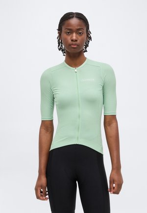 Noor inimene lühikeste patsidega, kes kannab helerohelise lühikeste varrukatega rattariietust ja musti rattapükse, seisab ühevärvilise tausta ees.