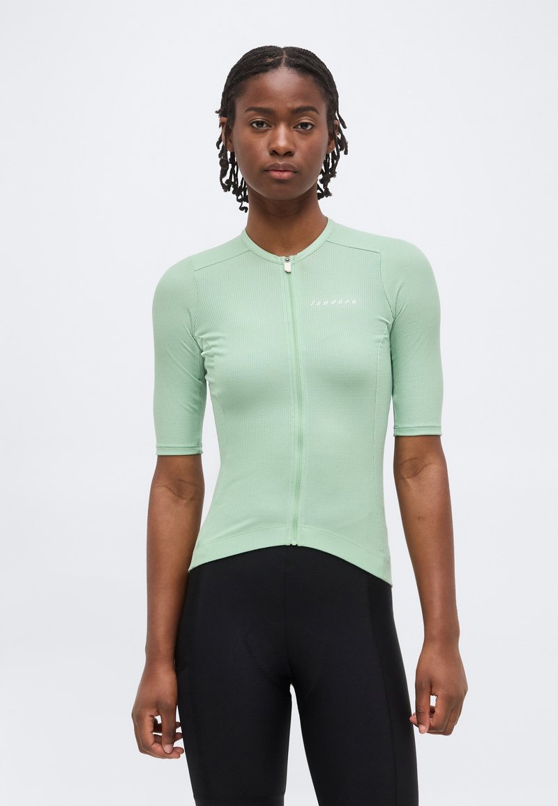 Νεαρό άτομο με κοντά πλεξούδες που φοράει ανοιχτοπράσινη κοντομάνικη κοντομάνικη φανέλα ποδηλασίας και μαύρα ποδηλατικά σορτς, στέκεται μπροστά σε ένα απλό φόντο.