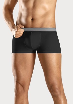 Oberkörper und Beine eines Mannes, der schwarze Boxershorts mit gestreiftem Bund trägt und den Bund mit der linken Hand leicht nach außen zieht.