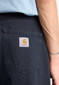 Sorte denimbukser med en baglomme, der har et hvidt mærke med et gult logo og rød tekst. Synlige syninger og en pæn afslutning.