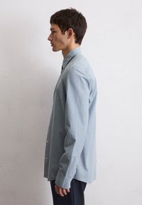 Chemise boutonnée en bleu clair avec des rayures blanches verticales ; elle dispose d'un col classique, de manches longues et de poignets à boutons. Tissu texturé.