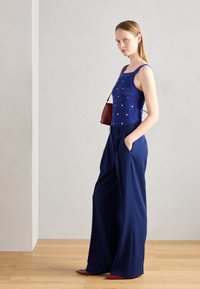 Top court en tricot bleu avec des points décoratifs, associé à un pantalon ample bleu marine. Les accessoires comprennent un petit sac à main bordeaux et des chaussures rouges.