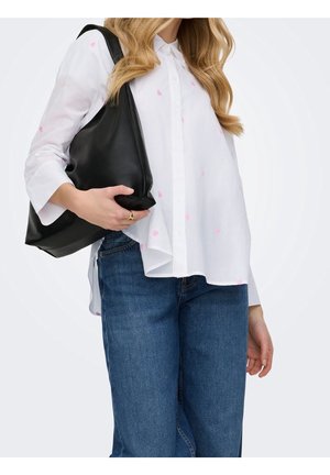 Femme portant une chemise blanche à boutons avec des cœurs roses et un jean bleu, tenant un sac à bandoulière noir avec sa main gauche.