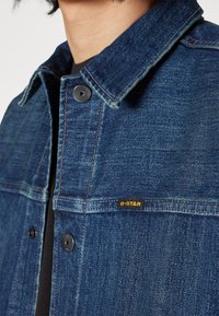 Jeansjacka i mörkblått med klassisk krage, med svarta knappar och en liten gul G-Star-etikett på vänster ficka.