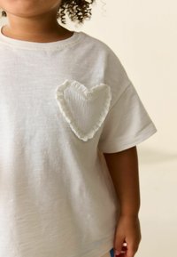 Weißes T-Shirt mit kurzen Ärmeln und einem strukturierten Herzpatch auf der linken Brust mit gerafften Kanten. Weicher Stoff mit entspannter Passform.