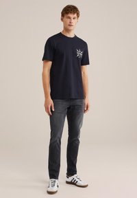 Tamno plava pamučna majica kratkih rukava s grafičkim dizajnom na lijevoj strani prsnog koša. Uparena s tamnim trapericama i bijelim tenisicama.