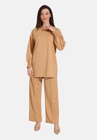 Beige tunika med samlede ærmer og matchende brede bukser, lavet af glat stof. Parret med lyse, spidse sko.