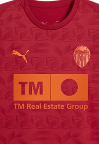 Röd atletisk tröja med ett strukturerat geometriskt mönster, en gul Puma-logotyp på bröstet och en orange sponsorlogotyp med "TM Real Estate Group."