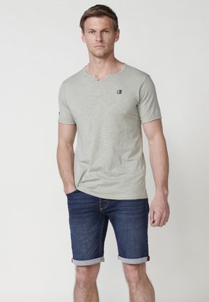 Camisa corta verde claro de manga corta con cuello redondo y un pequeño logo en el pecho, combinada con pantalones cortos de mezclilla azules oscuros con dobladillos enrollados.