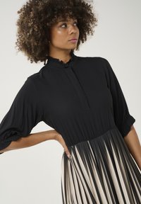 Čierne šaty s vysokým golierom, pufkovými rukávmi, sukňou s prekladom, ktorý prechádza z čiernej do krémovej farby, a mäkkou textúrou.