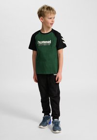 Grön och svart T-shirt med "hummel sportswear"-logotyp, korta ärmar med vita randiga accenter; kombinerad med svarta byxor och blå sneakers.