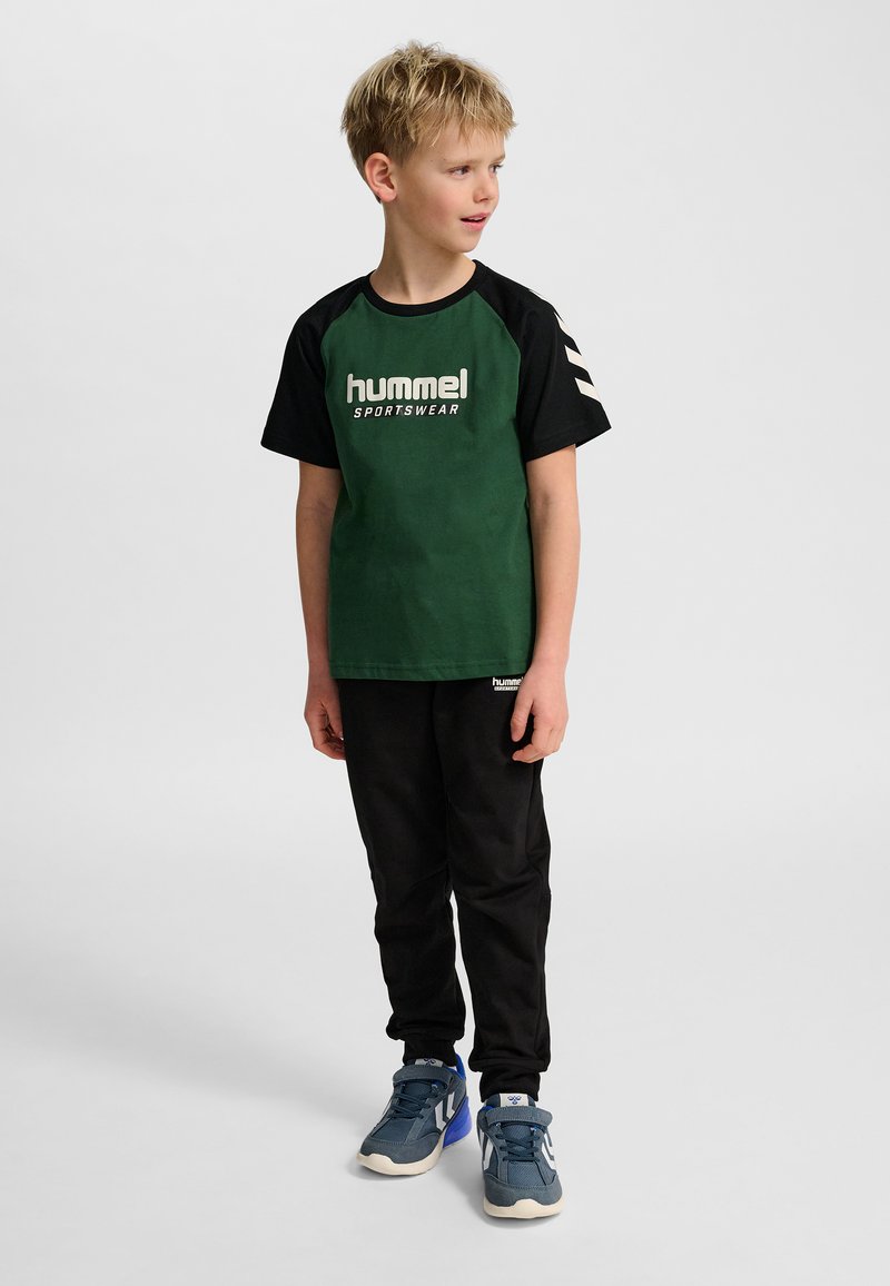 Grön och svart T-shirt med "hummel sportswear"-logotyp, korta ärmar med vita randiga accenter; kombinerad med svarta byxor och blå sneakers.