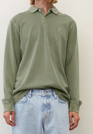 Henkilö, joka pukeutunut hillittyyn oliivinvihreään pitkähihainen poolopaitaan, joka on sullottu vaaleanpestyjen sinisten farkkujen sisään, yksinkertaisen taustan edessä.