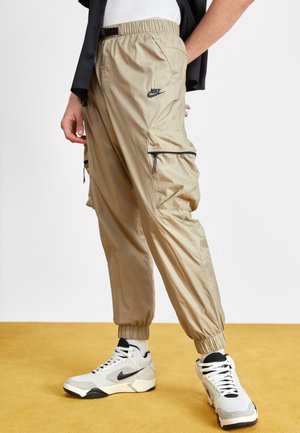 Beżowe spodnie typu cargo Nike z elastycznymi mankietami, czarne logo, noszone z białymi i szarymi sneakersami Nike z czarnym znakiem Swoosh.