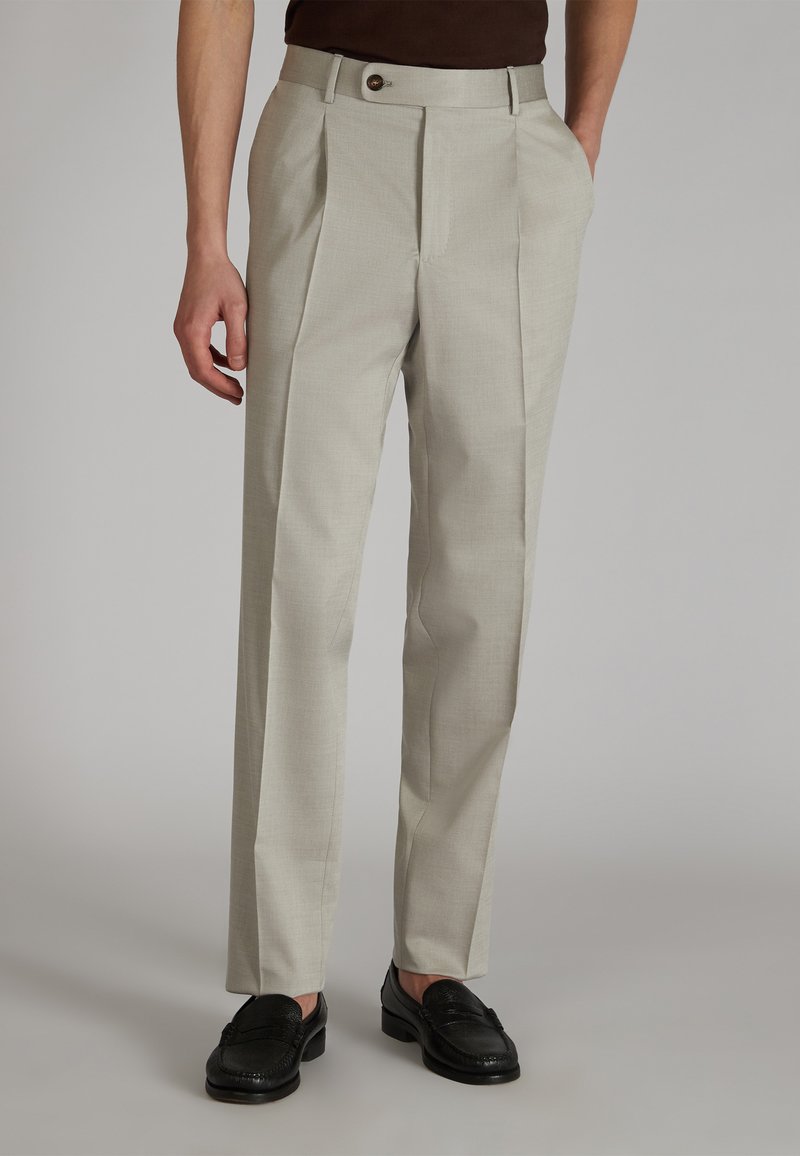 Beżowe spodnie o dopasowanym kroju z teksturowanym wykończeniem, z dwiema fałdami z przodu, kieszeniami bocznymi i pasem zapinanym na jeden guzik, zestawione z czarnymi butami.