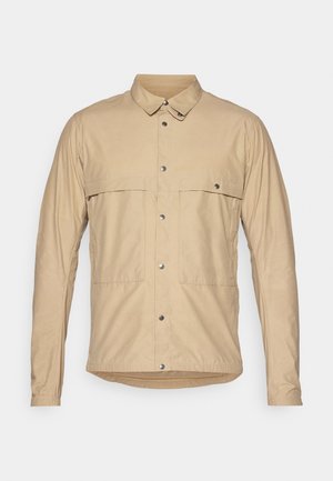 Beige, langermet skjorte med krage og knapper, som har to påsydde lommer, trykknapper og et glatt, lett stoff.