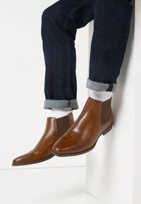 Brown leather chelsea boots with elastic side panels, pointed toe, and low heel, paired with dark denim and white ribbed socks.
