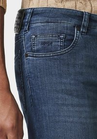 Jeans en denim dans un lavage foncé avec une coupe ajustée. Comprend une poche avant, un logo embossé sur la ceinture et des coutures visibles.