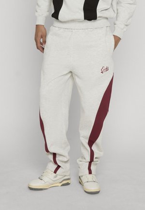 Personne portant un pantalon de survêtement gris clair avec des bandes latérales bordeaux, logo Ecko Unlimited sur la cuisse, associé à des baskets blanches.