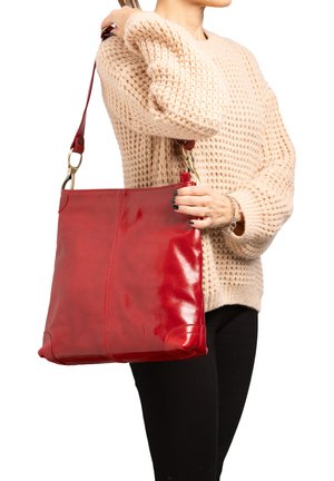 Vrouw met een grote glanzende rode leren schoudertas, gekleed in een beige gebreide trui en een zwarte broek, met de arm gebogen bij de elleboog.