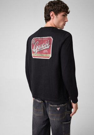 Joven con cabello rizado que lleva una camiseta negra de manga larga Guess y vaqueros oscuros, de pie y ligeramente girado de lado frente a un fondo liso.