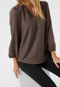 Blusa marrón texturizada con escote en V, mangas de tres cuartos y puños elásticos, combinada con pantalones negros ajustados. Diseño simple y versátil.