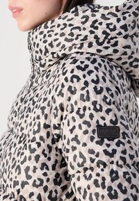 Nahaufnahme einer Frau, die eine beigefarbene Steppjacke mit Leopardenmuster und Kapuze trägt, wobei das Markenlabel "Barbour International" am Ärmel sichtbar ist.
