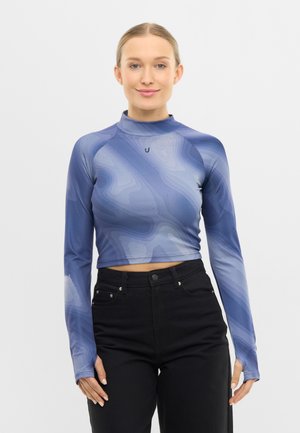 Nő, aki kék mintás, hosszú ujjú, felül rövidített felsőt visel fekete, magasderekú nadrággal, egyszerű világos háttér előtt áll.