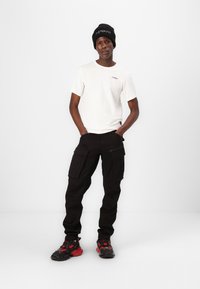 Vit t-shirt med kort ärm, svarta cargobyxor med flera fickor, och svart-röda sneakers med strukturerad sula och snörning. Svart mössa.