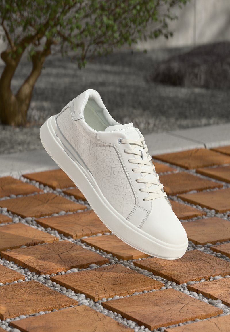 White sneaker with embossed pattern and laces, floating above a wooden tile and pebble pathway, with greenery and stone background.