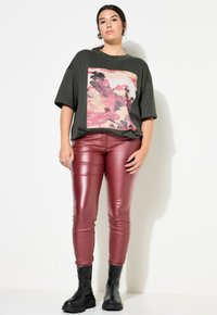 Olijfgroen oversized t-shirt met een levendige bergafdruk, gecombineerd met strakke, glanzende bordeauxrode neplederen broeken en zwarte chunky laarzen.