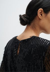 Top nero con paillettes, dal tessuto trasparente, caratterizzato da motivi a righe verticali e chiusura a occhiello sul retro. Accenti testurizzati e riflettenti in tutta la sua lunghezza.