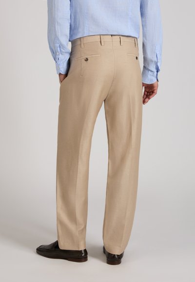 Homme portant un pantalon beige, une chemise à manches longues bleu clair et des chaussures marron foncé, debout avec les mains dans les poches, vue de dos.