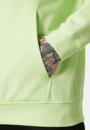 Dłoń z kwiecistym tatuażem częściowo wsunięta do przedniej kieszeni jasnobrzozowego swetra.