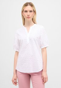 Hvid broderet bluse med V-udskæring og korte puffede ærmer; har blomsterformede hulmønstre og en let tekstur.
