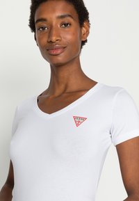 Camiseta blanca de cuello en V con mangas cortas, que presenta un logotipo triangular rojo en el pecho izquierdo. Textura suave, ajuste casual.