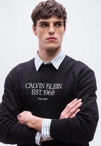 Νεαρός άνδρας με σταυρωμένα τα χέρια, φορούν μαύρο φούτερ Calvin Klein πάνω από ένα μπλε και λευκό ριγέ πουκάμισο με κουμπιά, κοιτάζοντας στο πλάι.