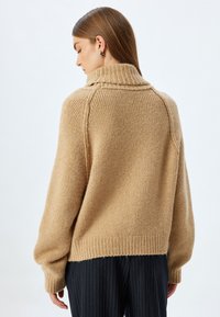 Maglione beige con colletto alto a coste, tessuto a maglia texture, spalle scese e polsini oversize, abbinato a pantaloni a righe sottili.