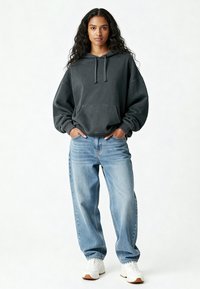 Giovane donna in piedi con le mani in tasca, indossa un ampio felpa scura, jeans larghi di un blu chiaro e sneaker bianche su uno sfondo bianco.