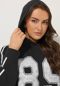 Felpa nera con un grande logo bianco consumato "89". Presenta un cappuccio con coulisse e righe bianche sui lati delle maniche. Tessuto morbido.