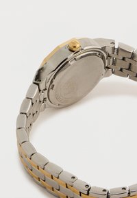 Μεταλλικό λουράκι ρολογιού από ασήμι και χρυσό με στρογγυλό πίσω μέρος κάσας προς τα επάνω, εμφανίζοντας την πόρπη και το κορώνα σε λευκό φόντο.