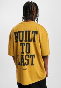 Gul oversized t-skjorte med svart tekst som sier "BUILT TO LAST" på baksiden, laget av bomull med korte ermer og rund hals.