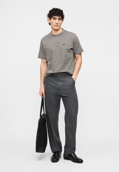 Νεαρός άνδρας που φοράει κοντομάνικο πουκάμισο με σχέδια, γκρι παντελόνι, μαύρα παπούτσια, κρατώντας μια μαύρη τσάντα tote, στέκεται μπροστά σε απλό φόντο.