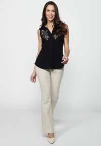 Schwarze ärmellose Bluse mit floralem Stickdetail, kombiniert mit hellbeigen Schlaghosen und beigen Schuhen, vor einem weißen Hintergrund.